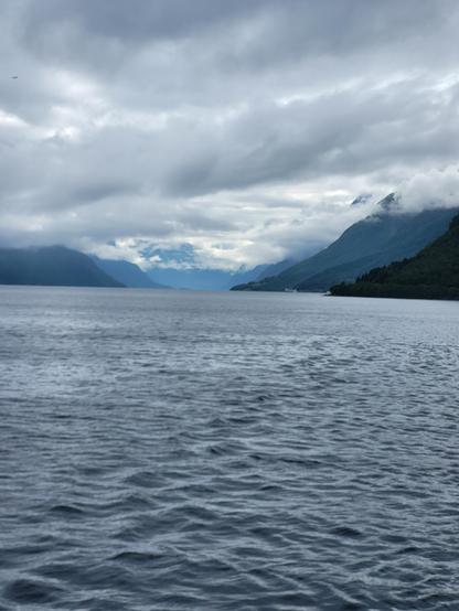 Im Vordergrund ist Wasser von einem See in Norwegen zu sehen. auf der rechten Seite ist ein Berg der mit vielen Bäumen bewaldet ist. Dieser ist dunkel grün. Auf der linken Seite ist eine Bergkette diese ist in Blautönen gehalten. Der Himmel ist komplett mit Wolken verhangen. Die Wolken sind gau und hängen sehr tief bis in die Berge hinein.
