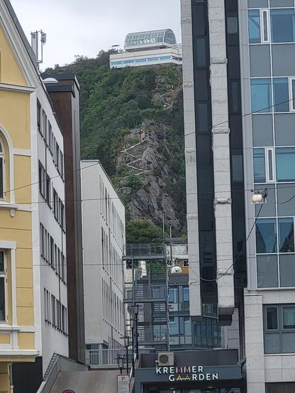Links und rechts und im mittleren unteren Teil sind Häuser. Zwischen den Häusern ist ein Berg mit einer Treppe und einer Aussichtsplattform. Der Himmel ist komplett grau.