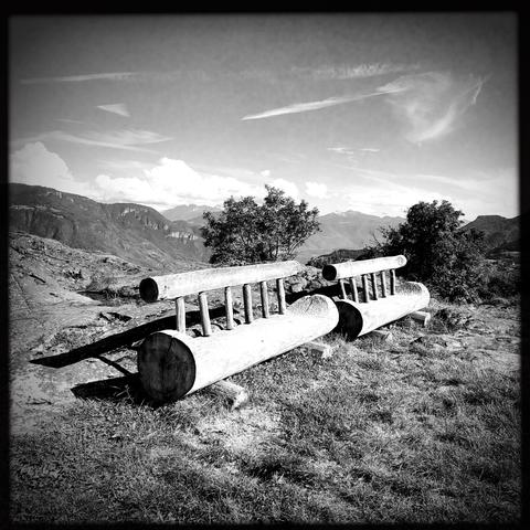 Due panchine con schienale realizzate con due tronchi su di un prato con le cime delle montagne sullo sfondo