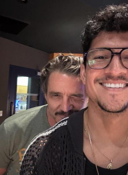 Photo showing Danny Ramirez wearing glasses and standing in the foreground with Pedro Pascal peeking over his shoulder.