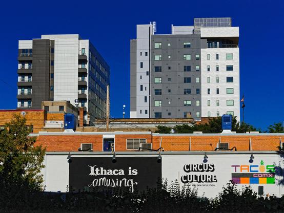 In front of a dark blue and prefectly blue sky find two gray and white apartment buildings,  the one on the left a bit smaller than the one on the right but both about the same height if you take perspective into account.  Below those a row of light brown brick buildings and a white wall with murals that say: Ithaca is amusing, CIRCUS CULTURE and TACOS COMX with some vegetation in silhoutte in the foreground of that.