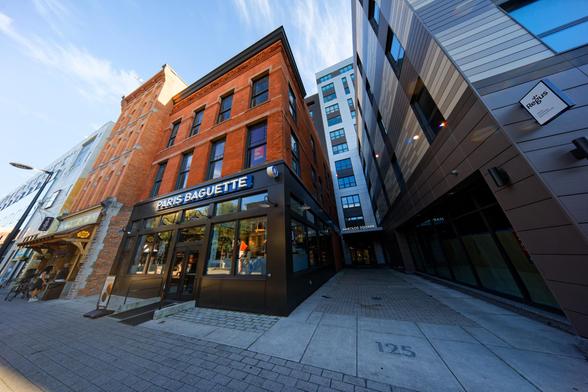 A unique wide perspective on an urban scene,  on the 1/3 line to the right is a narrow path with the number 125 carved into the concrete walkway and a view to a column of 5 clusters of windows rising up from an entrance with the sign HAROLDS SQUARE.  To the right of that is a building with a dark grey floor level and alternating white and grey stripes rising up from that,  to the left an old brick building that has a PARIS BAGUETTE on the first floor that looks like an urban McDonald's from the…