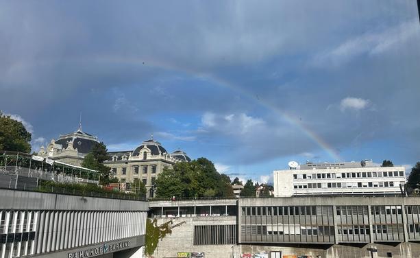Blick aus dem Fenster auf einen relativ wolkigen Himmel mit blauen Flicken. Ein Regenbogen spannt sich über den Gebäuden der Berner Uni über dem Bahnhof.