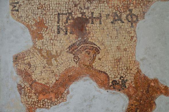 Mosaic of Aphrodite in warm, reddish colours. Only the head, shoulders, and arms remain. She wears a bracelet and earrings. She wears her hair in an up-do with either a band or crown.