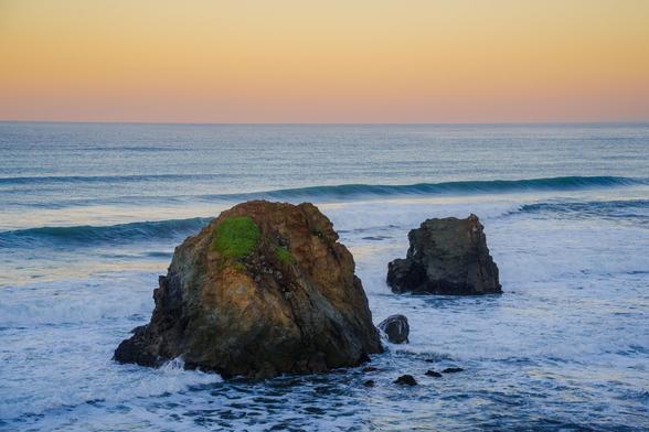 Two large rocks in the ocean with gentle waves and a colorful sunset in the background.