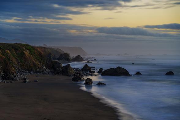 A beach scene with rocky cliffs, misty waves, and scattered rocks along the shore under a cloudy sky.