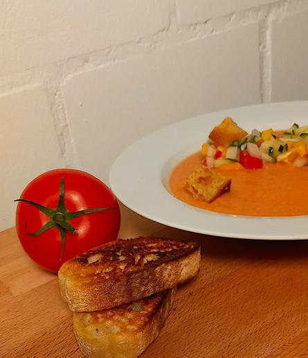 Foto zeigt rechts im Bild einen weißen Teller mit Gazpacho, links davon eine Tomate und zwei scheiben getoastetes Weißbrot.
