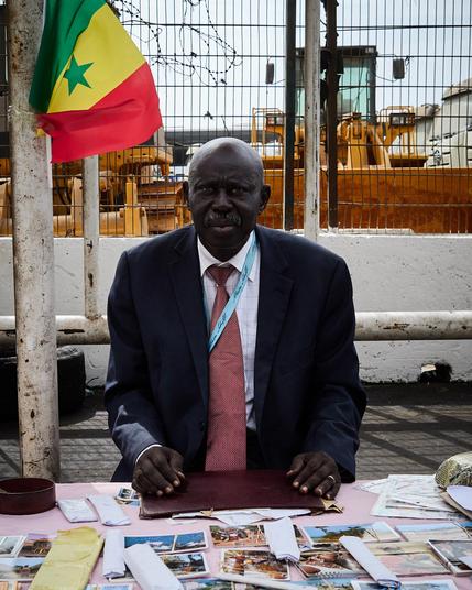 A man in a suit and tie sits at a table covered with postcards and letters. He is outdoors, against a backdrop of construction equipment. A flag of Senegal is visible in the background.