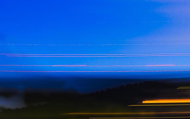 An abstract view of the late dusk sky caused by panning the camera across the scene during a long exposure. The sky above is deep blue, with the ground below darker. Hints of trees can be seen below, from the ends of the pan. There are also orange lights from houses and buildings below. Above there are several horizontal red lines from the lights on communication towers – one is blinking slowly on and off, creating incomplete trails. Then above that is the star Antares, which is fairly faint close the horizon, but you can see its brightness and colour changing due to atmospheric turbulence. There is another fainter star trail above it. There are a number of small point-like white dots presumably from airplanes with flashing lights. 