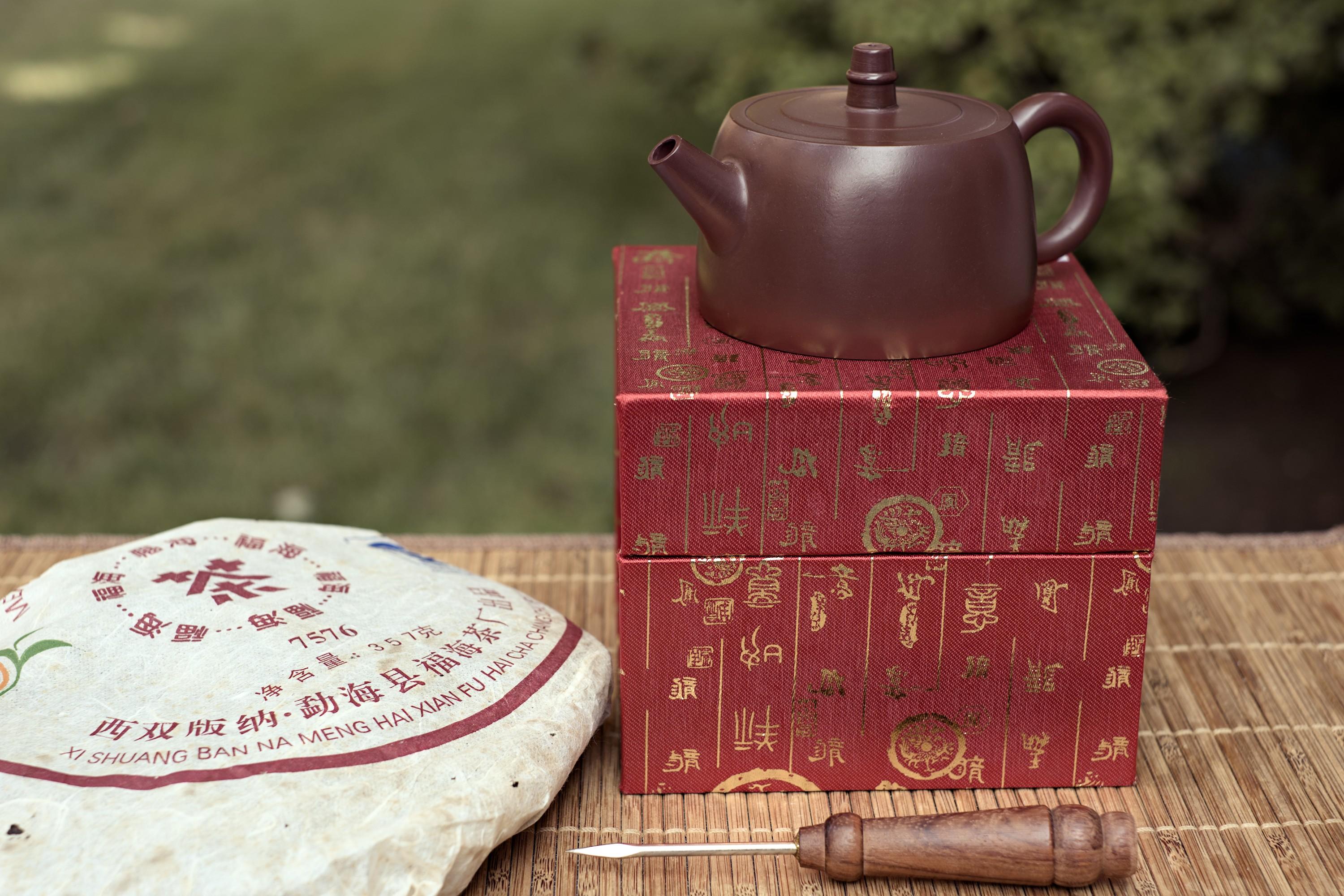 Small dark brown clay teapot with a wide flat base slightly tapering to the top. No adornments to speak of. It is standing on a red box inscribed in golden Chinese characters. A wrapped cake of puerh tea and a puerh knife lay in front of it. A non-descript grass lawn is in the background. The picture has a slight sepia tint to it.