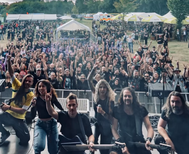 Das Video zeigt eine lebhafte Live-Auftrittsszene auf einem Musikfestival. Im Vordergrund steht die Band DARKK auf der Bühne, bestehend aus mehreren Musikern, die Gitarren und andere Instrumente spielen. Die Musiker tragen schwarze Kleidung, und einige von ihnen machen die berühmte "Rock on"-Geste mit ihren Händen. Im Hintergrund ist eine große, begeisterte Menge von Fans zu sehen, die die Bühne mit erhobenen Armen und Gesichtern voller Energie betrachten. Viele der Fans tragen ebenfalls schwar…