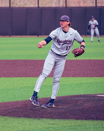 Baseball pitcher stands with his left foot on the ground and his right foot off the ground pointing at and close to his left wearing brown shoes, black socks, grey pants and jersey with a green undershirty and blue billed cap with a P on it,  the jersey reads Quakers 17 and another Penn player can be seen in the outfield behind him.