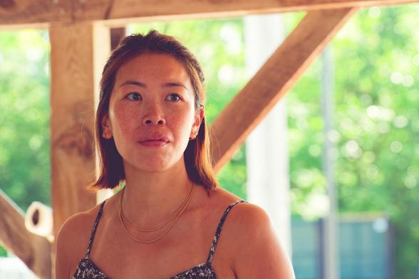 Asian woman with hair just above her bare shoulders,  a necklace and top with thin straps looks to the right (her left) with closed lips,  behind her can be seen the wood braces of the farmer's market pavillion and past that bright green leaves with sparkles of light passing between them
