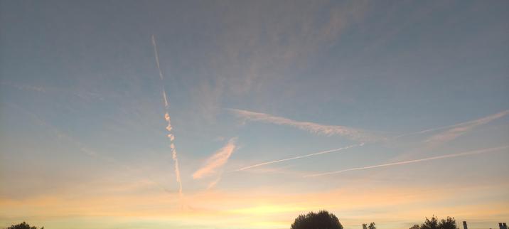 Ciel de l’aube aux teintes pastel, passant du jaune doré à un bleu clair doux. Plusieurs traînées d’avion traversent la toile, dont une verticale en pointillés, comme des perles suspendues. En bas, les silhouettes sombres des arbres encadrent la scène.