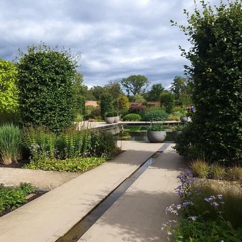 A view of the central water feature in the Paradise garden at Bridgewater. A narrow water  rill leads to a square pond where waterlillies float on the surface. The rill runs through light coloured gravel with additional paths leading off at right angles. The flower beds are planted with a mixture of flowers and structural shrubs.  In the distance the horizon is filled with mature trees.