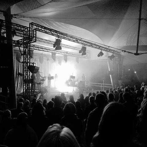 Zwart-witfoto van een concert van Eefje de Visser in het Goffertpark Theater. Het podium is fel verlicht van achteren, waardoor de muzikanten in silhouet te zien zijn. Voor het podium staat een groot publiek in de schaduw, onder een grote overkapping.