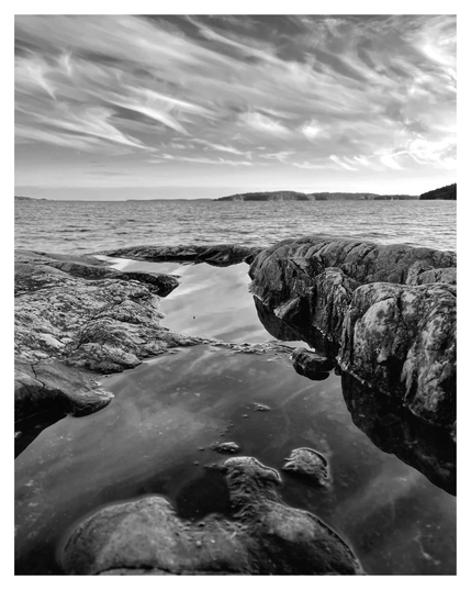 Black-and-white photograph of a rocky shoreline with tidal pools in the foreground, leading the eye toward the calm sea and distant islands on the horizon. Wispy, streaked clouds stretch dramatically across the sky, reflected faintly in the still water between the rocks.