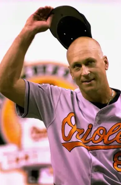 Cal Ripken Jr., wearing a gray Baltimore Orioles jersey with orange script, smiles warmly as he lifts his cap in acknowledgment—likely during a celebratory moment on the field. His shaved head glows under stadium lights, and the blurred background features a large commemorative emblem, suggesting this photo was taken during or after his historic 2,131st consecutive game. His gesture is humble, but the moment feels monumental—like a working-class superhero bowing without ever breaking.