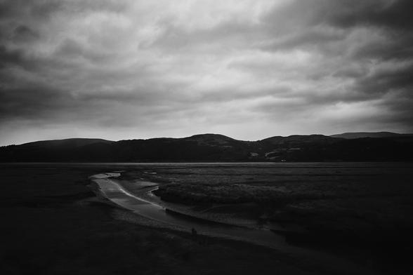 The image captures a moody scene of a tidal river winding through the landscape. The sky is filled with dark, heavy clouds and the hills in the background create a sense of depth. The river reflects the overcast sky, and the surrounding area is mostly flat with patches of grass, adding a serene yet dramatic atmosphere.
