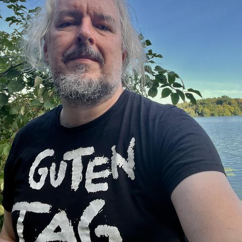 Selfie (50-jähriger mit Bart und grauen Haaren) vor See mit einem schwarzen T-Shirt, auf dem in großen freundlichen Buchstaben "Guten Tag" steht.