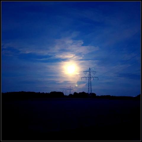 A huge moon rising over a field at nightfall. With a row of transmission towers beginning at the top right of the picture and moving towards the bottom left and into the distance.