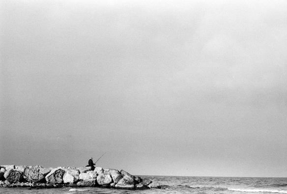 This is a black-and-white photograph with a strong minimalist composition.

In the lower left part of the image, a lone person is sitting on large rocks by the sea, fishing with a rod. The figure appears small and solitary against the vast expanse of the sky and ocean. The rocks form a breakwater or jetty, extending slightly into the water.

The upper two-thirds of the image is dominated by an open, grainy sky, giving a sense of emptiness and calm. The horizon line is low, placing emphasis on the vast sky rather than the water. The sea in the bottom right corner looks slightly wavy, contrasting with the stillness of the fisher.

Overall, the photo evokes a feeling of solitude, patience, and contemplation, enhanced by the monochrome tones and wide negative space.