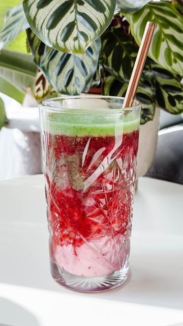 A tall etched glass filled with a layered berry smoothie: green top layer, a deep red middle layer with visible berry fragments, and a pale pink bottom layer, with a copper straw angled in from the right. A leafy plant with variegated green leaves sits in the background.