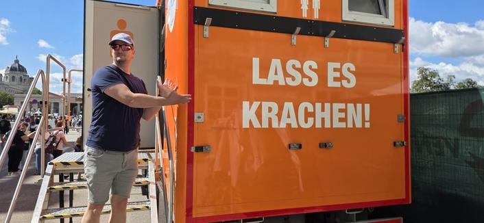 Ich vor einer mobile Toilette am Heldenplatz. Darauf befindet sich die Aufschrift "Lass es krachen!". Ich deute auf den Schriftzug.