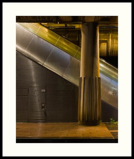 colour - Side view of an escalator leading to an elevated tram platform. Taken on an early, rainy, and still dark morning. Most of the scene is illuminated by lightly coloured orange nitrate lamps on the ground floor. Some white light is coming from the platform above and accentuates certain areas in the scene.