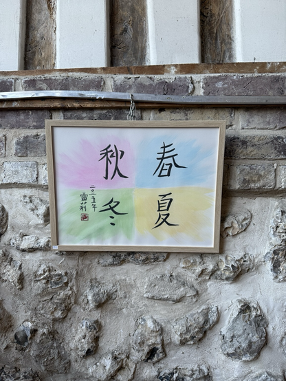 Cadre accroché sur un mur de pierre, représentant les quatre saisons écrites en calligraphie chinoise : 春 (printemps), 夏 (été), 秋 (automne), 冬 (hiver), sur un fond coloré pastel en dégradés rose, jaune, vert et bleu. 
