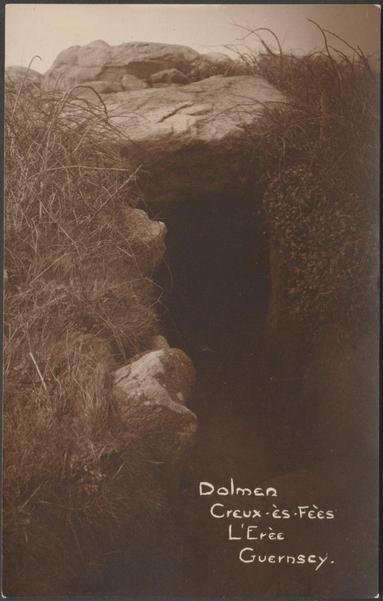 A vintage sepia postcard showing the entrance of Le Creux-ès-Fèes or Le Creux ès Faïes dolmen, a passage grave or chambered tomb near St Peter Port on Guernsey in the Channel Islands. A narrow passage leads to a bottle-shaped chamber roofed with a series of large, chunky capstones.