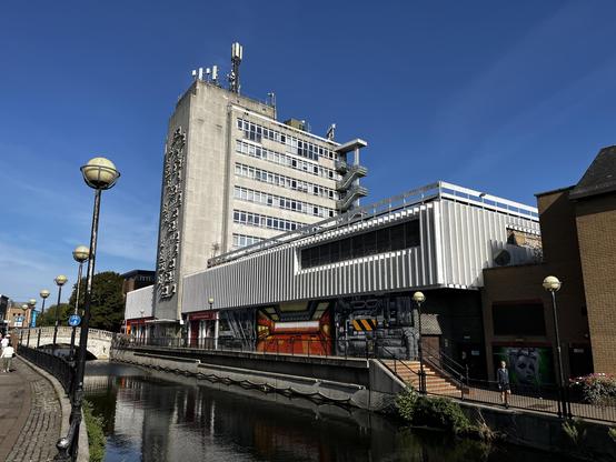 Ground-level shot of a mid-height 1960's concrete building, with a large mass at ground level and the next few storeys extending higher in a more narrow mass. On the lowest level there are shops, as well as some graffiti. Above, the next two storeys are hidden behind concrete panels, with the level above featuring a visible fence around it, suggesting a roof terrace. The actual concrete block is fairly typical for its age, but interestingly featuring a concrete relief on one size, and an external staircase on the other.