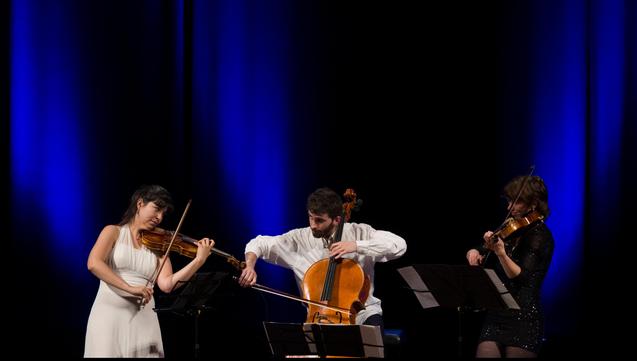 De gauche à droite : Lia, Marc et Coline en train de jouer respectivement du violon, violoncelle et alto. Lia en blanc, Marc en noir et blanc, Coline en noir à paillettes. Derrière, des rideaux noirs éclairés de lumières bleues roi.