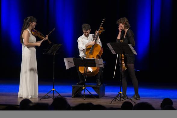 De gauche à droite : Lia qui vient de s'arrêter de jouer, souriant. Marc rit et remet sa corde de do en place sur son violoncelle. Coline rit et se cache la bouche de la main droite.