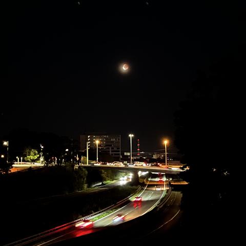 Nacht, Autos auf einer Schnellstraße (rote und weiße Lichter, zu Strichen verzogen), oben drüber klein der Mond. Dunkle Mondscheibe, links unten hell, aber leider sehr pixelig-verschwommen.