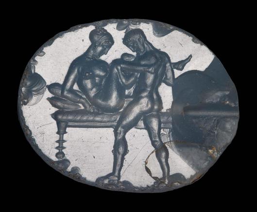 Intaglio. Pierced for stringing (horizontal). Motif oriented horizontally. A woman, seated on a cushioned bed, has intercourse with a male partner. Her face and legs are seen in profile, while her torso is twisted toward the viewer in a viewpoint somewhere between three-quarters and frontal. The woman wears a beaded necklace, a bracelet on her right arm, earrings, and thin slippers. Her hair is bound up in a scarf, with a few locks of hair escaping at her forehead and crown. She has a long nose…