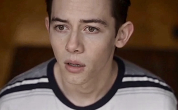 A white teenage boy looking worried. He’s wearing a white and grey T-shirt with black stripes on the shoulders.