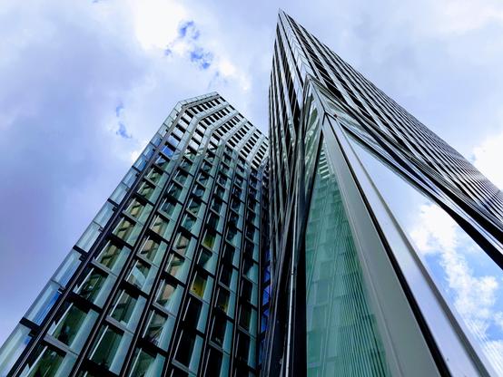 Bildbeschreibung von KI:
Das Foto zeigt die tanzenden Türme in Hamburg, zwei markante Hochhäuser an der Reeperbahn. Die Aufnahme ist von unten nach oben fotografiert, sodass die Türme besonders hoch und imposant wirken. Beide Gebäude bestehen fast vollständig aus Glas und Stahl. Ihre Fassaden sind von vielen rechteckigen Fenstern durchzogen, die in einem leichten Versatz angeordnet sind und so einen unregelmäßigen, dynamischen Eindruck erzeugen. Der besondere Effekt der Architektur liegt darin, dass die Türme leicht gegeneinander verdreht sind, wodurch es aussieht, als würden sie sich in einer tänzerischen Bewegung zueinander neigen. Die Glasfassaden spiegeln den Himmel, in dem Wolken zu sehen sind, teils hell, teils grau, was dem Bild Tiefe verleiht. Die Linienführung der Gebäude zieht den Blick stark in die Höhe, sodass man das Gefühl bekommt, mitten in der Szenerie nach oben zu schauen. Die Architektur wirkt modern, kühn und kraftvoll und spiegelt den charakteristischen Stil dieses Wahrzeichens von Hamburg wider.
