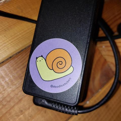 A black laptop charger on a wooden floor. The charger has a small round sticker on it. The sticker has a purple background, with a small, cute, cartoon snail, which has a green body with a tiny wee face, and a brown shell. The name @doodlesbyben is written below the snail in a curve.