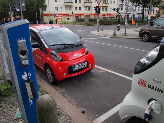 Two Citroën C-Zero compact electric vehicles (rebranded Mitsubishi i-MiEVs) from the Deutsche Bahn car-sharing scheme are parked next to a blue charging station. In the background is a view of a Berlin street.