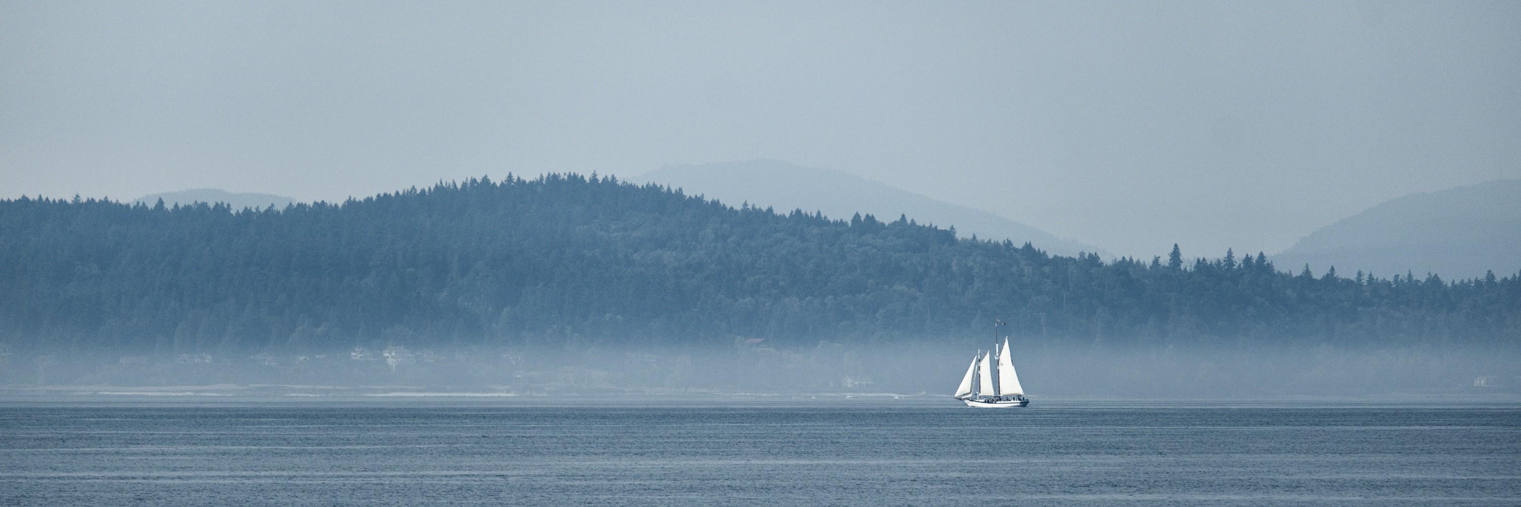 A view over Elliott Bay from Seattle waterfront with a clear expanse of water, a shining white sailboat far away and a wooded island in the background. The photo is colored in a cool blue hue.