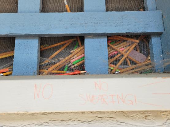 Daytime photo of the exterior of a screen window barred with painted wooden bars, behind which a few dozen yellow pencils and colored pencils loosely rest haphazardly in a pile. On the white bottom exterior window border, someone has written "NO NO SWEARING!" with a reddish-orange colored pencil.