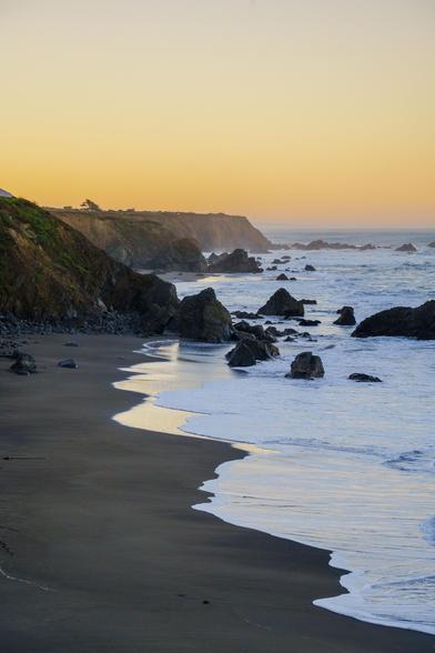 A tranquil sunset view of the beach, where soft waves meet the sandy shore, surrounded by rocky cliffs.