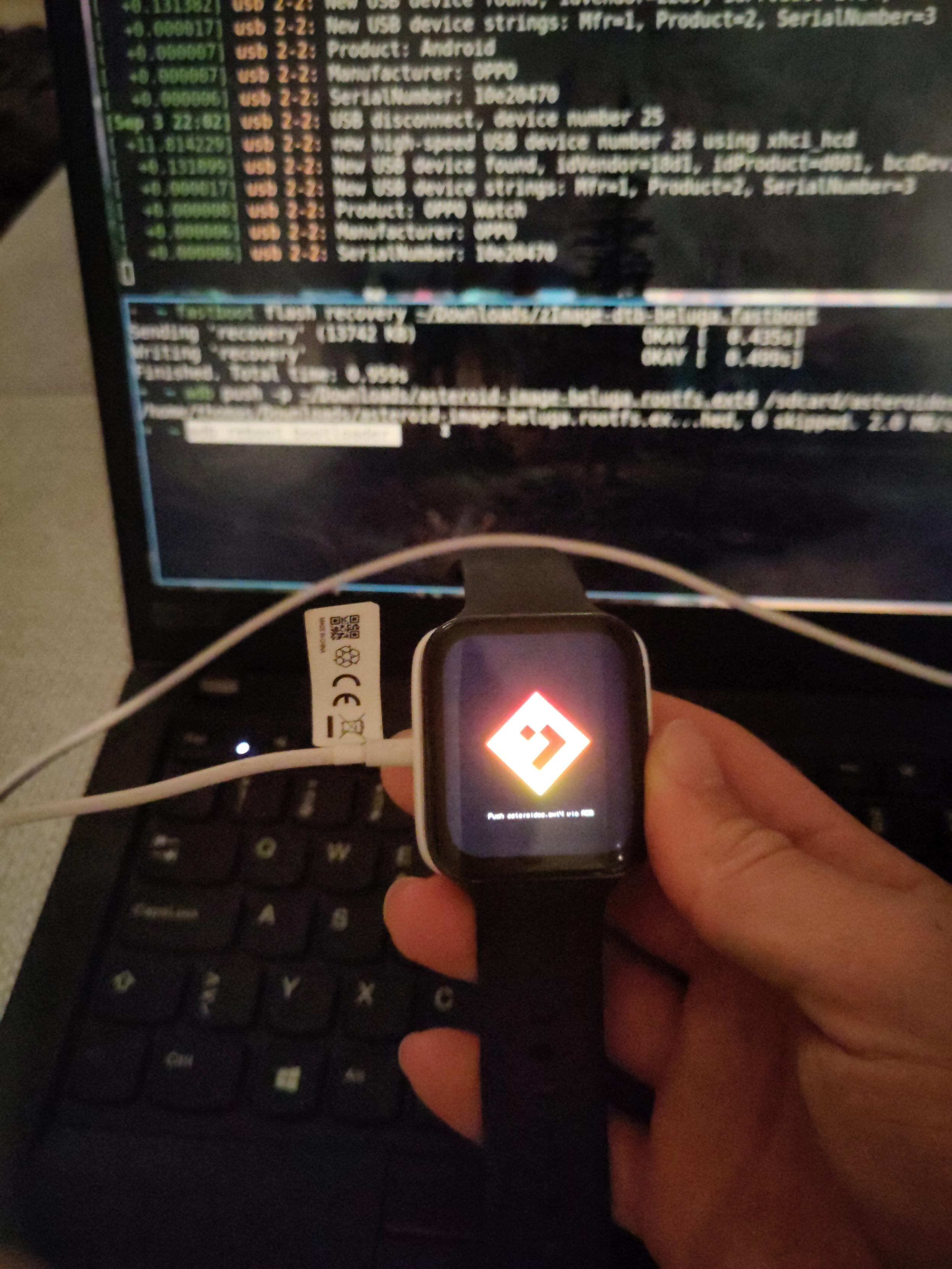 Holding the Oppo watch which displays the AsteroidOS logo with a message "Push asteroidos.ext4 via ADB". In the background my Thinkpad can be seen with two terminals, displaying dmesg kernel logs and abd/fastboot commanda respectively.
