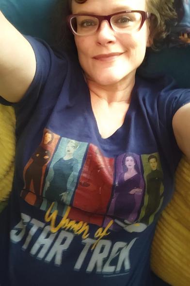 Waist-up selfie of a middle-aged white woman (me) with short, dark, curly hair and glasses. I'm wearing a dark blue shirt that says "Women of Star Trek" (with "Women of" in a messy yellow printed font and " Star Trek" jn the TOS font). Above the words are five colored panels, each depicting a head-to-knees portrait of a woman in Star Trek: an orange panel with Janeway, a light blue panel with Seven, red with Uhura, purple with Troi, and green with Dax.