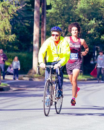 In the dead center is a cyclist on a road bike wearing a blue helmet and high vis yellow vest,  behind him is a white man wearing a red tank top with blue lines says Cornell with a blue bib that says 354 in white and pink shoes with his feet entirely off the ground with some telephone poles foliage and a few onlookers behind.