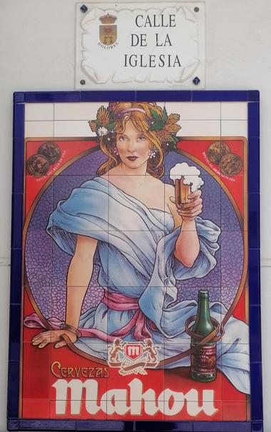 Ein Jugendstilbild einer Dame mit Kranz in den Locken und einem Bier in der Hand. Unten das Logo  von Mahou. Darüber hängt das Strassenschild Kirchenstrasse (auf spanisch).