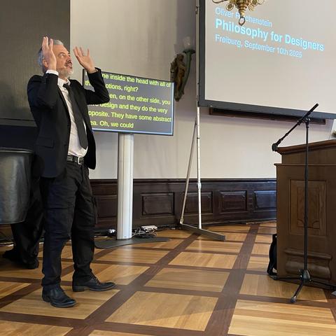 Oliver on stage, wearing a suit and raising his arms, with the title slide of the talk next to him that reads Philosophy for Designers