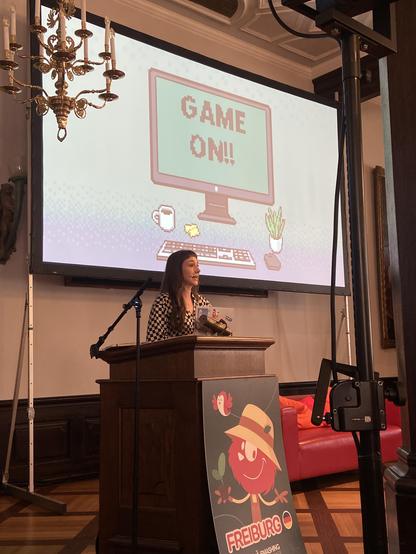 Cassie on stage with the title slide of the talk behind her. It reads Game On!