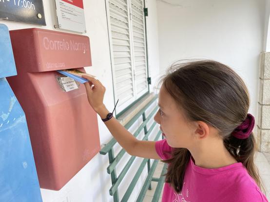 A young girl in a pink shirt posts a postcard into a red mailbox labeled “Correio Normal” in Portugal.